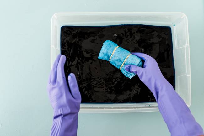 A person with blue gloves putting blue banded cloth into black colored water box.
