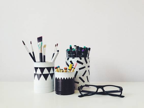 Tin cans dressed up into a patterned pencil cups filled with pencils, pens and brushes and a spectacle placed near it.
