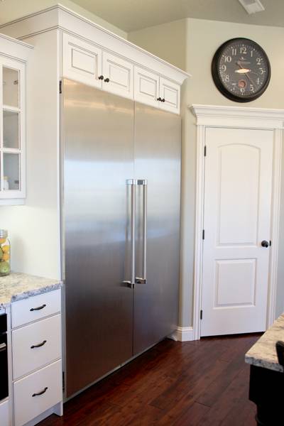 Corner of a nice white kitchen with a huge double door built in stainless steel refrigerator and a clock above a door.