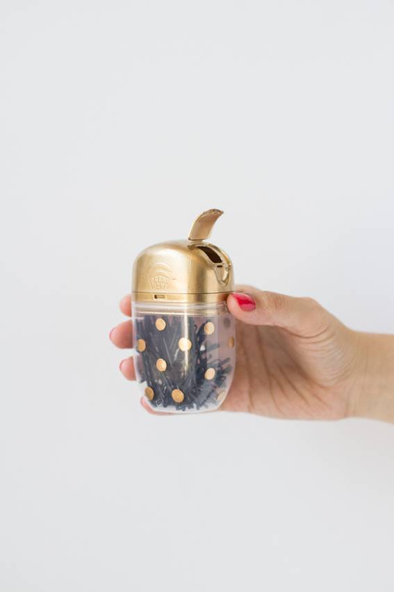 A hand with red nailpolish is holding a small clear container with gold polka dots and a gold top.