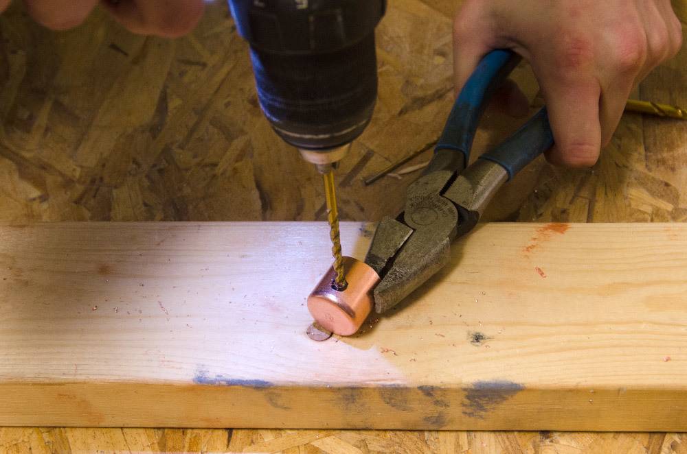 A man holds a copper cap with a pair of pliers and uses a drill to drill a hole in it.