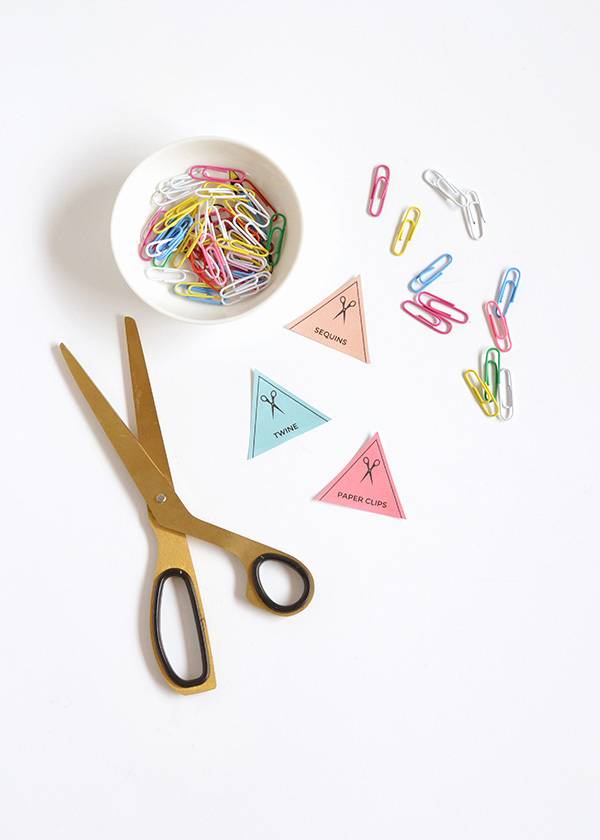 Scissors and paper triangles are on a white table next to a bowl of paperclips.