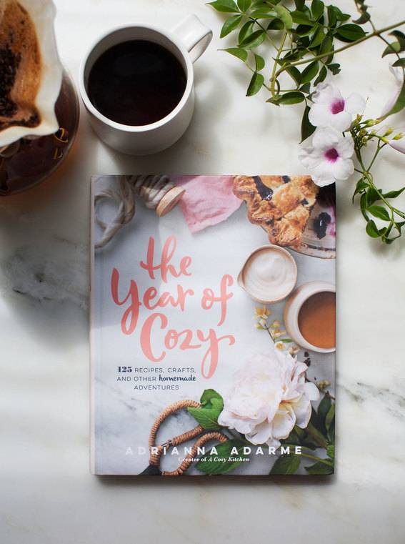 A book titled, the year of cozy, is laying on a marbled background next to a cup of black coffe and two white flowers sitting on some green twigs.