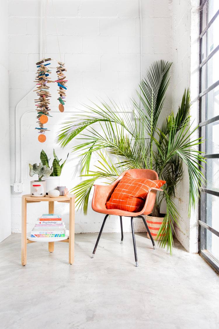 An orange retro chair with an orange pillow on it in front of a potted palm tree in the corner of a white room.