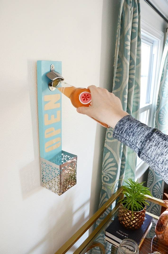 A person is taking the cap off a bottle of soda using a wall-mounted bottle opener.
