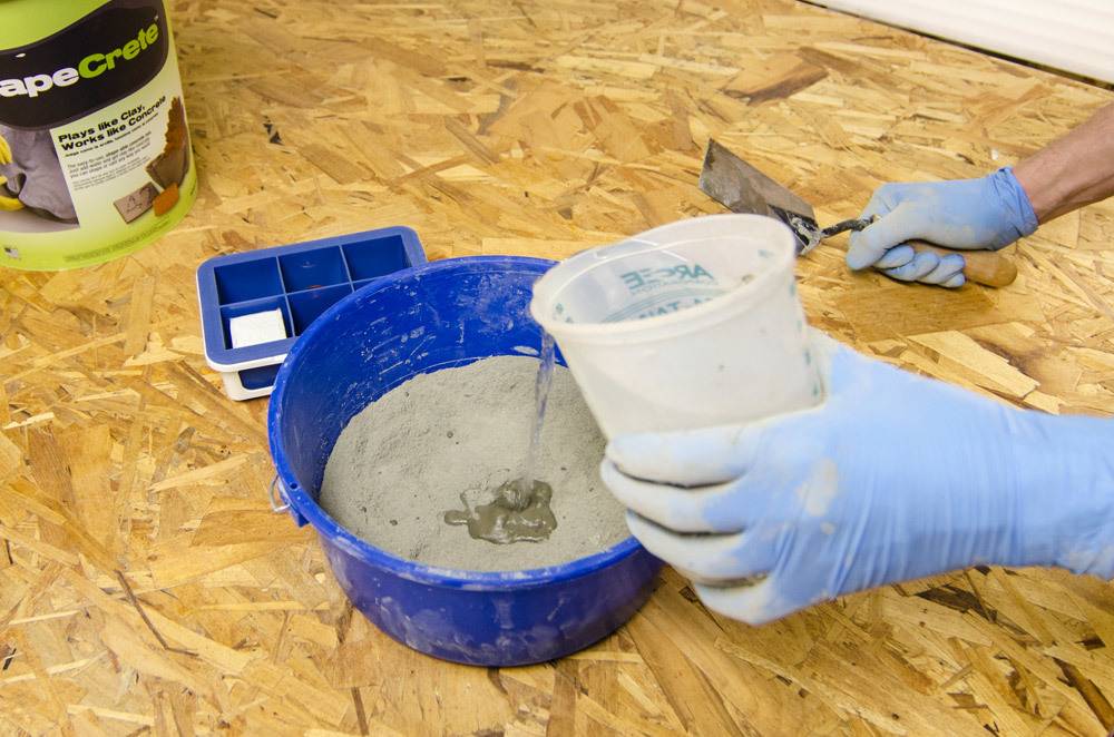 A person with light blue gloves mixes some cement in a blue container.