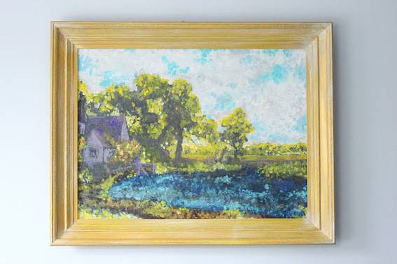 A tan framed painting of a purple country house by some trees behind a lake on a cloudy day.