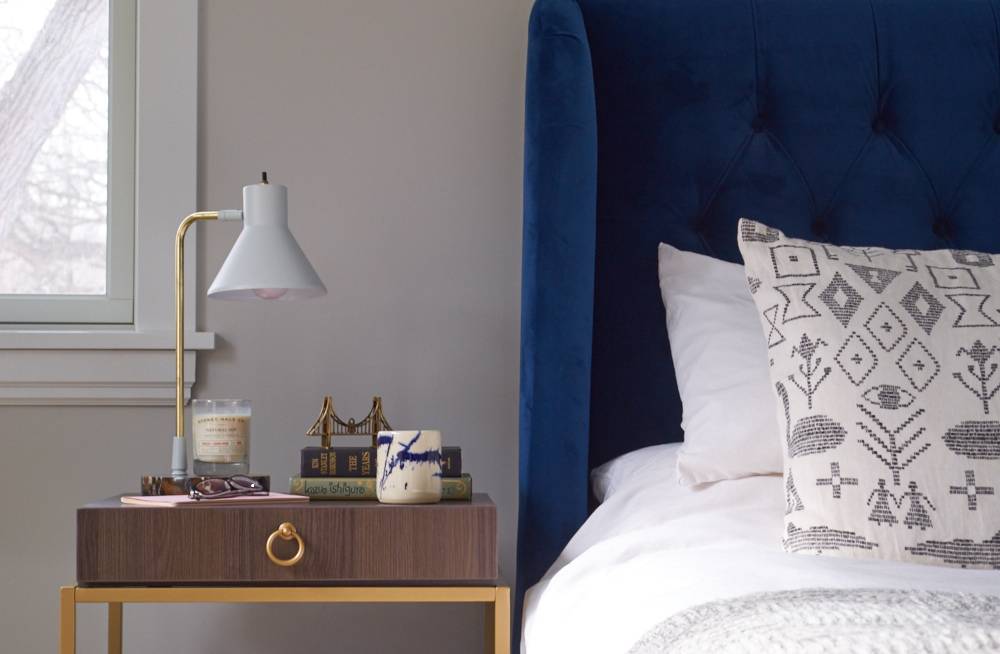 A bedroom has a dark blue bed header and white blankets next to a small end table with a lamp on it.