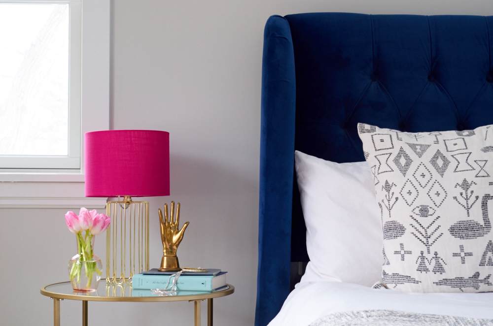 Nightstand with pink table lamp, flower vase, hand shaped gold color idol and book beside the bed.