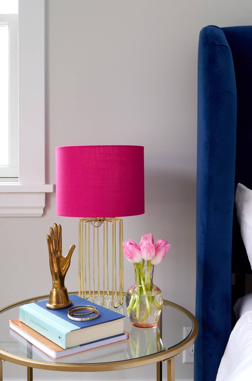 A lamp with a pink shade is sitting on a bedside table with books and trinkets.