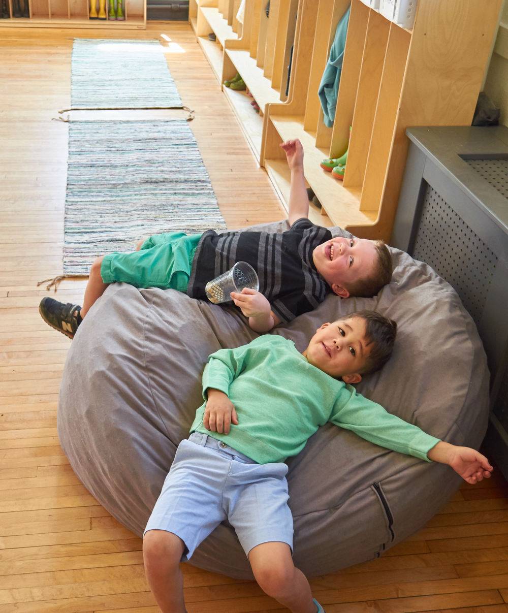 Two young boys lie on a grey bean bag chair in a room with wooden floors.