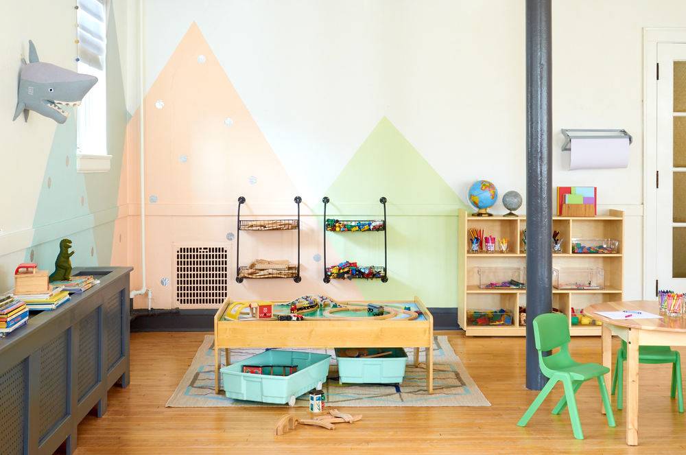A children's room, with a small table and green chairs next to a low play table with a sharks head on the wall and pink and green mountains painted on the wall.