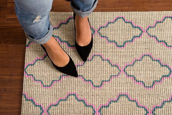 A woman's legs from the knee down wearing jeans and black pumps is standing on a tan carpet with blue and pink design outlines on it.