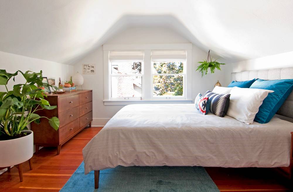 A bed with white sheets and blue and white pillows stands in a room with wooden floors and sloping ceilings.