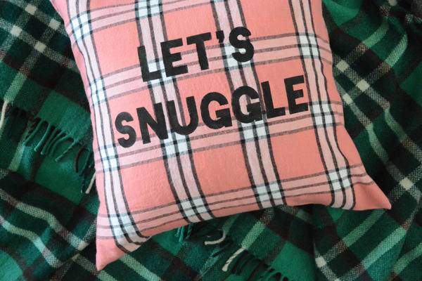 A plaid pink, white and black pillow that says "let's snuggle" atop a green, black and white plaid flannel blanket.