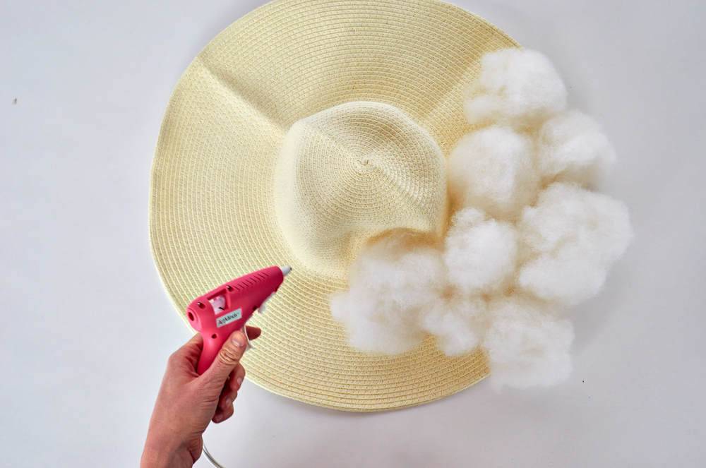 A woman is using a pink glue gun to attach cotton balls to the brim of a beige-colored hat.
