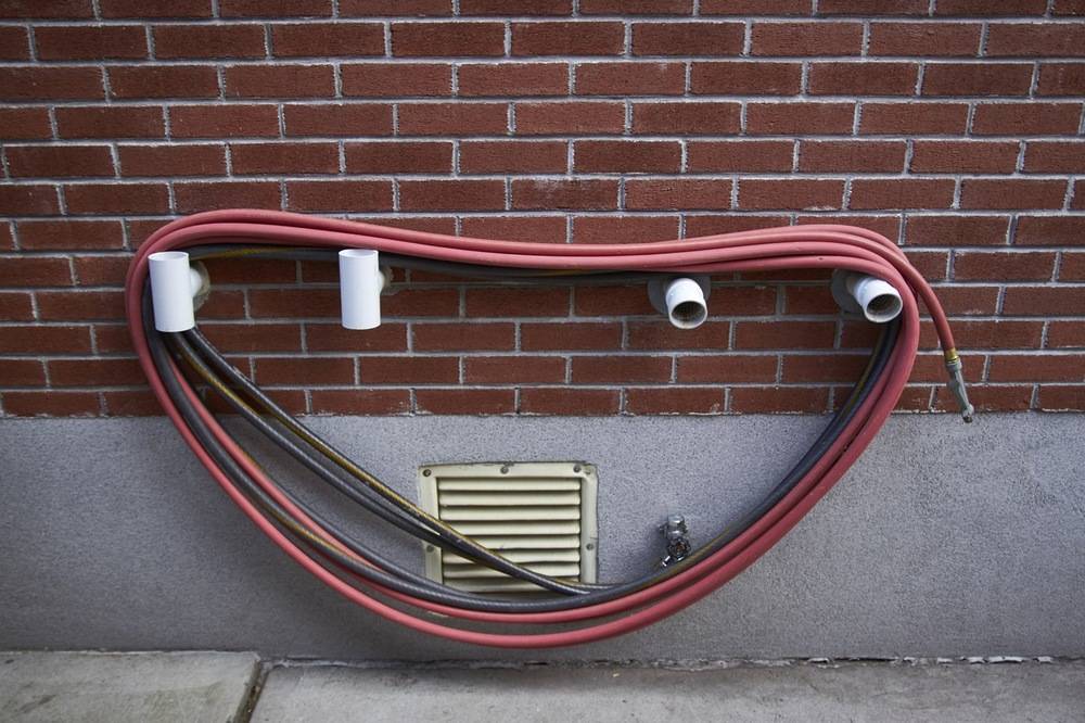 A red hose looped around PVC pipes on the side of a brick building.