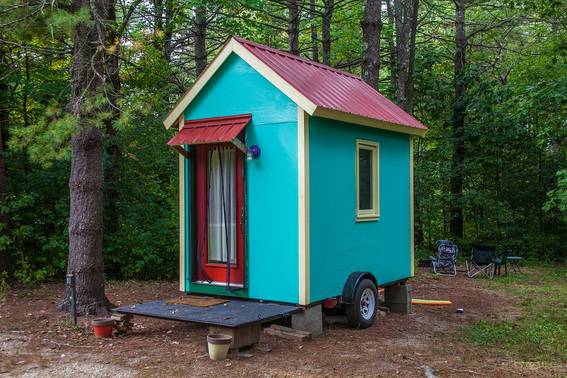 A very small, multi-colored house is mounted on blocks and also has tires attached, sitting in a wooded area.