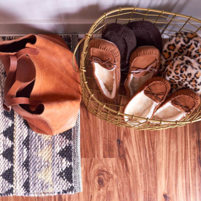 A wire basket of moccasin slippers next to a plain brown purse sitting on a grey rug that's laying on a wooden floor.