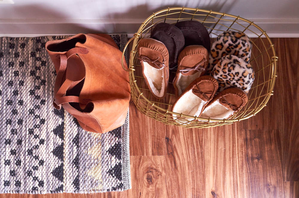 A wire basket of moccasin slippers next to a plain brown purse sitting on a grey rug that's laying on a wooden floor.