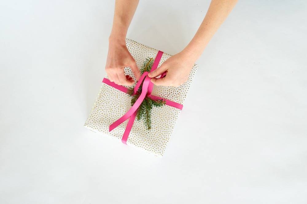 A pair of hands about to finish tying a pink ribbon around a small box that's wrapped in dotted wrapping paper.