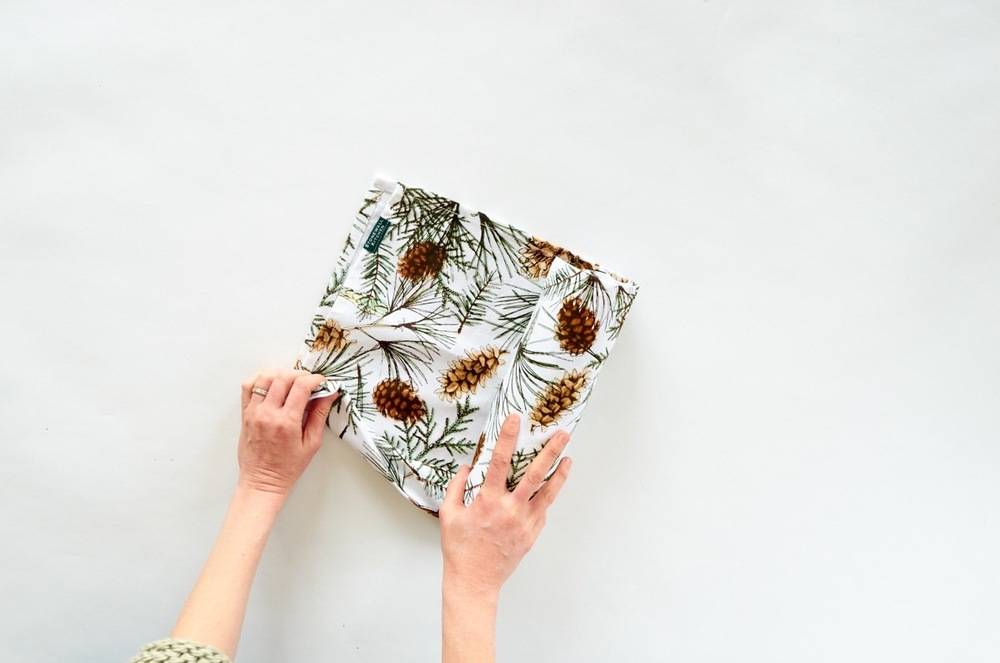 Two women's hands holding a folded piece of cloth decorated with brown pine cones and green tree twigs.