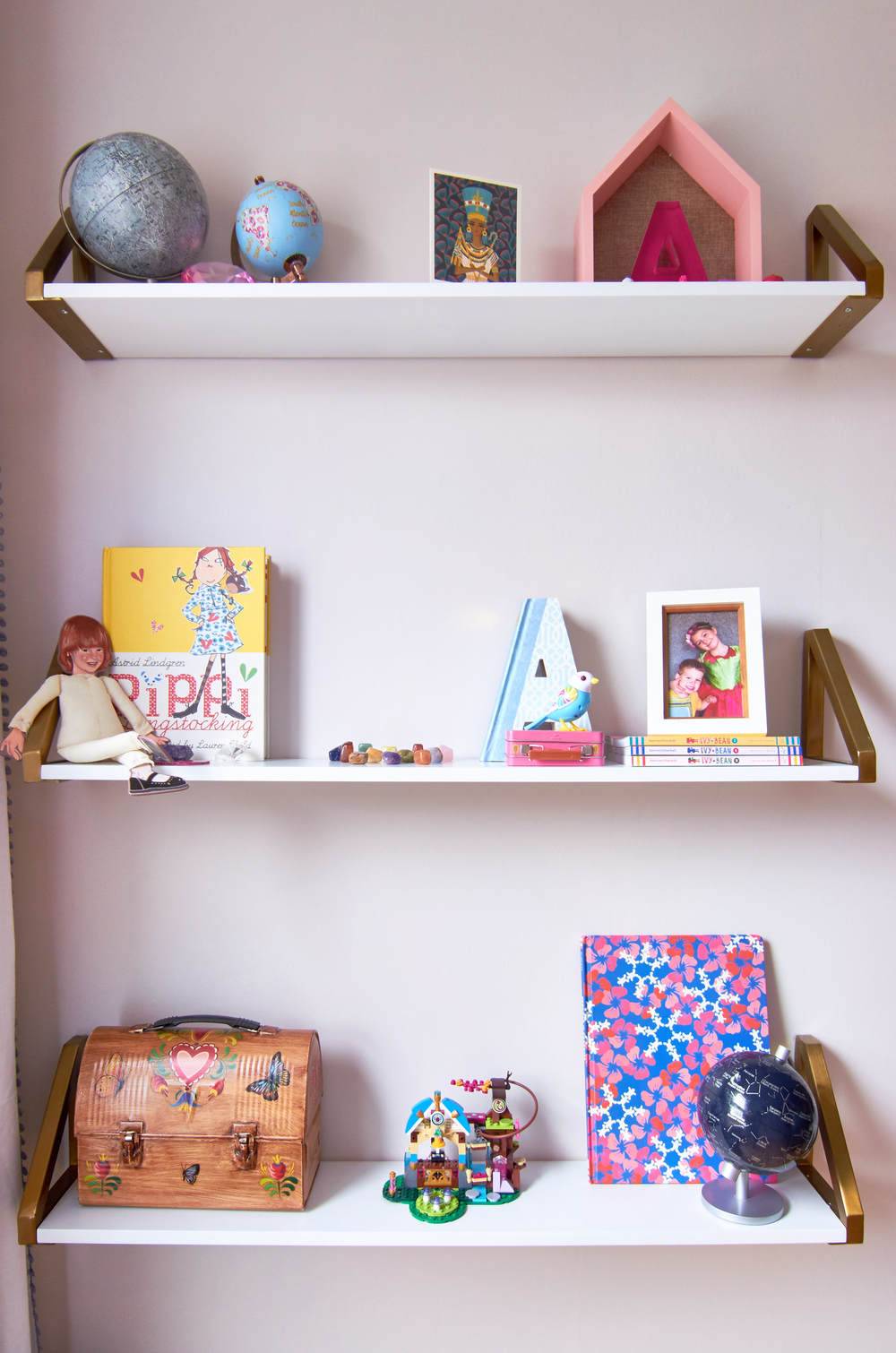 Three white floating shelves with a glove, a pink hous, a yellow book, a brown lunch pail and a pink and blue notebook on them.