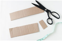 A piece of cardboard cut into three pieces, next to a pair of scissors and measuring tape.