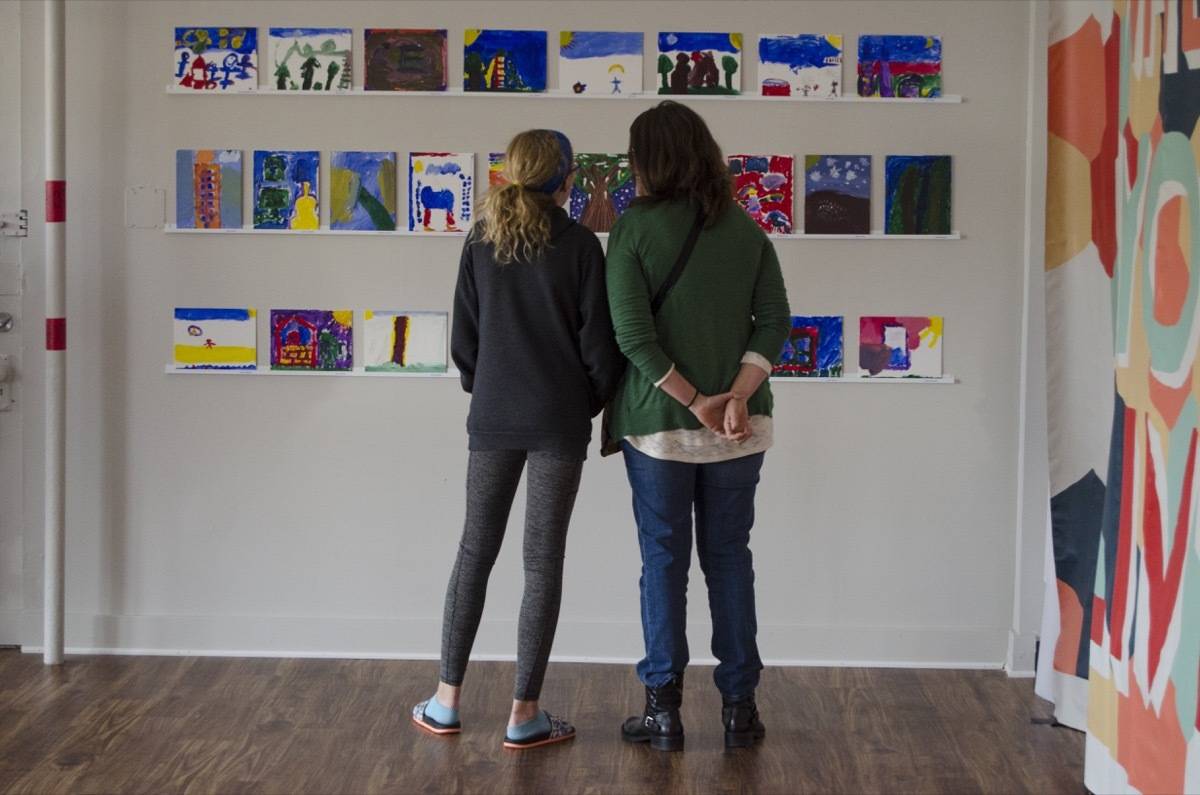 A mother and a daughter looking at three rows of artwork on a white wall.