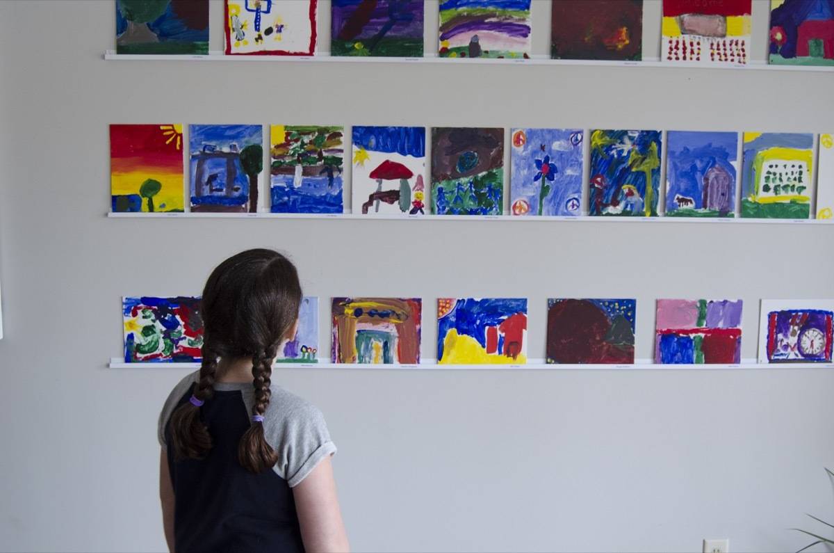 Young girl looking at several pieces of children's artwork displayed neatly on a wall.