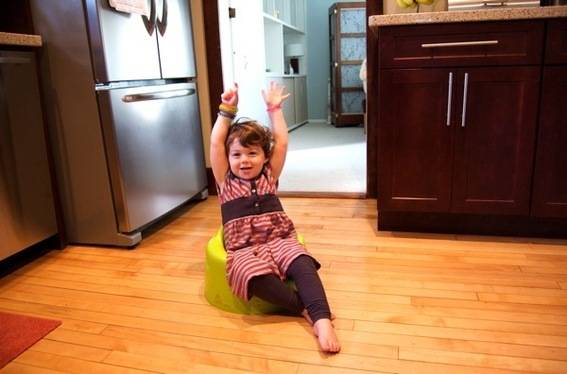 A young girl with her hands in the air and six fingers pointing up, she is sittng on a yellow booster seat in a kitchen.