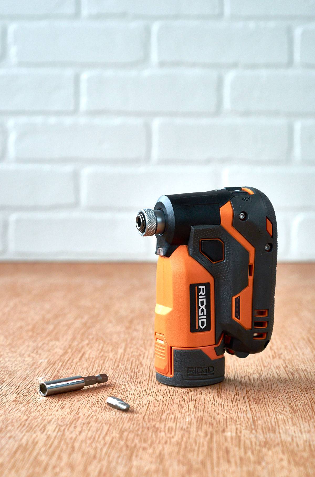 A drill with two different drill bits on the floor next to a brick wall.