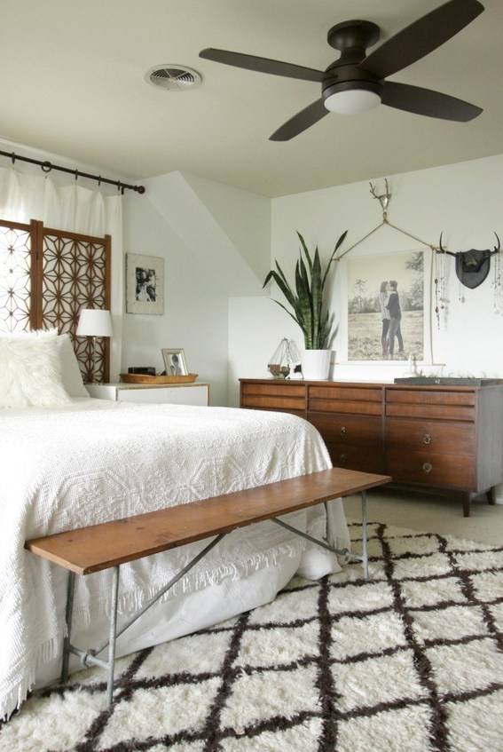 A bed sits on a rug with a criss cross pattern in a room with a ceiling fan.
