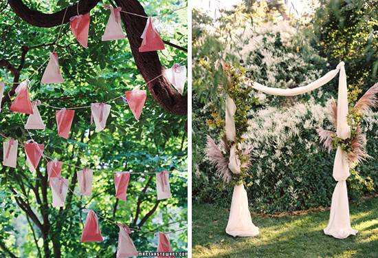 Triangular flags on a string hanging from a tree next to a ribbon archway by a bush.