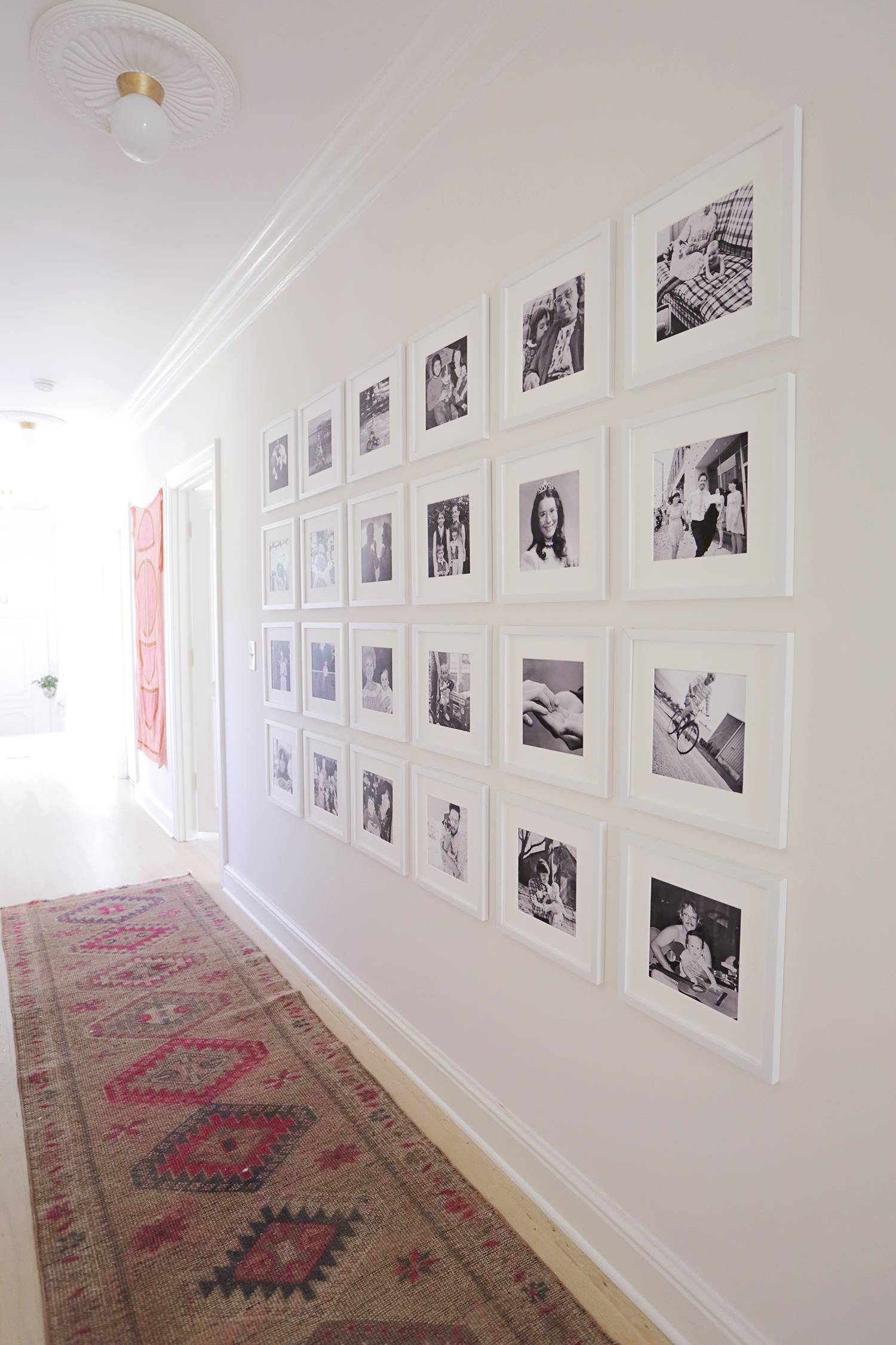 A hallway with a red and brown diamond decorated runner rug and four rows of six, black and white pictures on the wall.