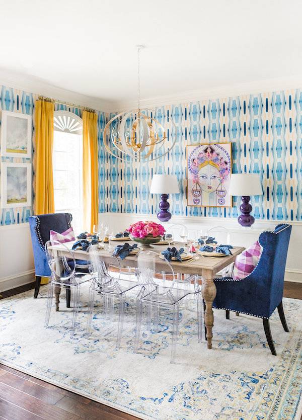 A dining room table with three see through chairs on the side of the table and a blue chair on each end in front of a window with yellow shades and blue and white wallpaper.