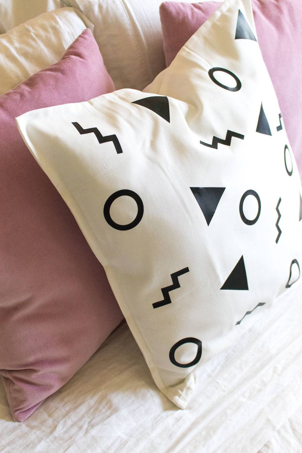 A white pillow with black triangles, circles and squigels sits in front of two pink pillows on a bed.