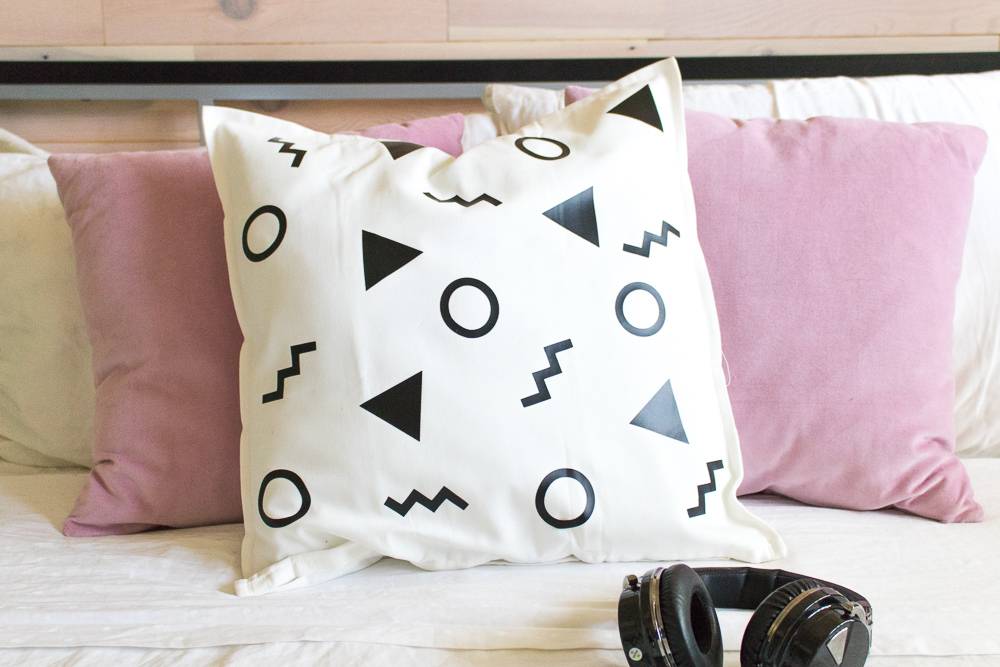 A bed has two pink pillows and one white one with designs on it.