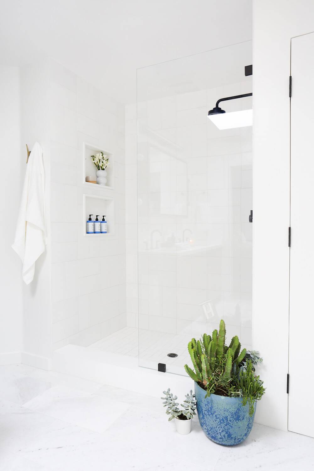 A white shower with a white towel hanging on the wall and two plants, one in a blue planter and one in a tiny white planter next to the shower.