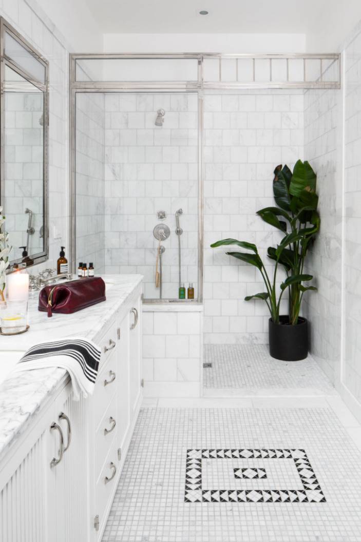 A white bathroom with a large green plant in the shower, a red mens shaving bag on the marble countertop and a black square tile design in the middle of the floor.