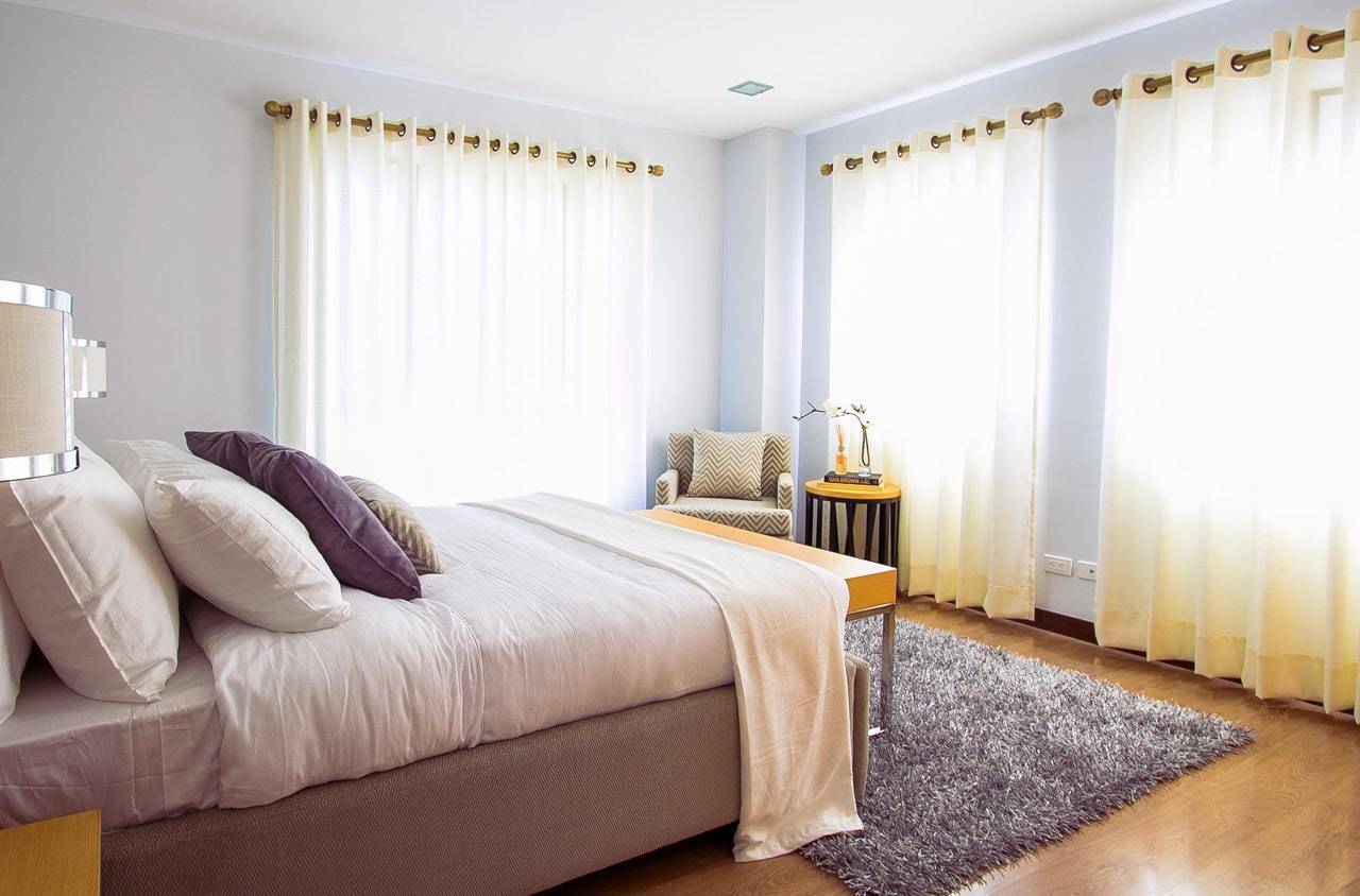 The sun shines through white curtains ina. bedroom with a large bed and end table.