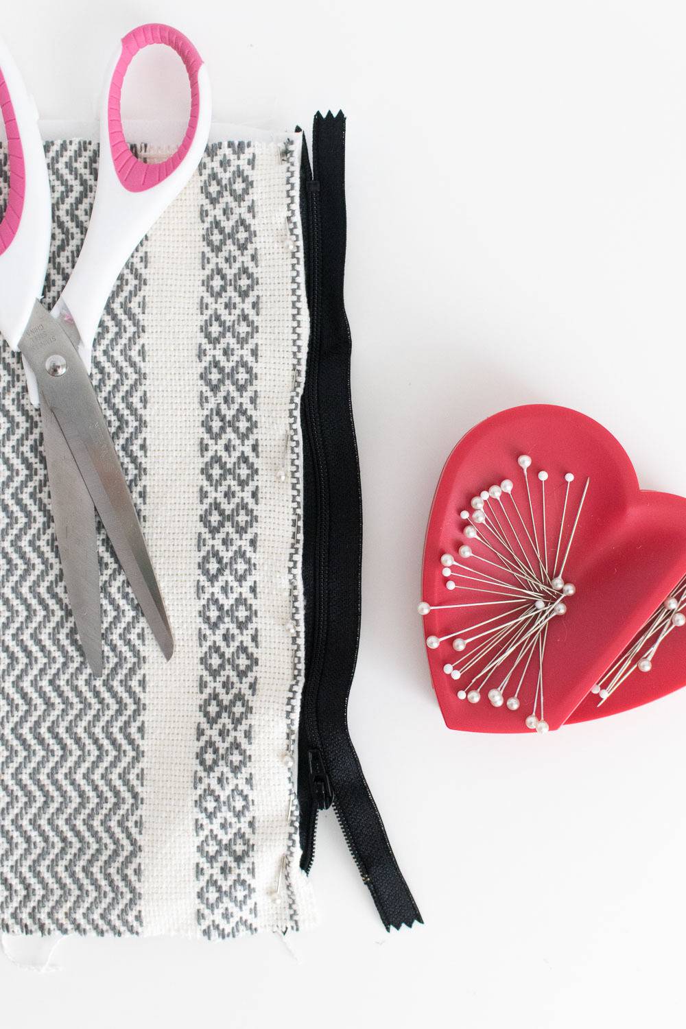 A pair of pink and white scissors, a grey and white piece of cloth, a black strip of ribbon and a red heart pin holder.
