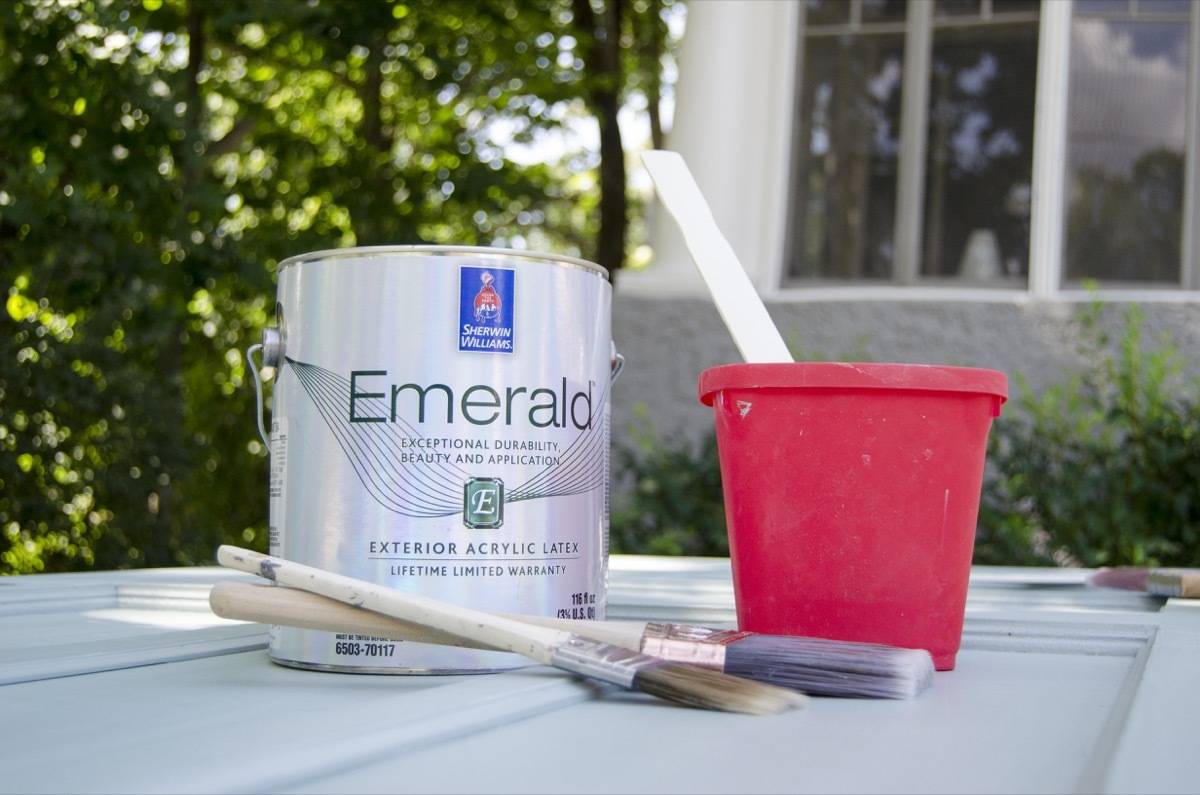 A can of latex paint sitting next to a pair of paintbrushes and a small red plastic bucket.