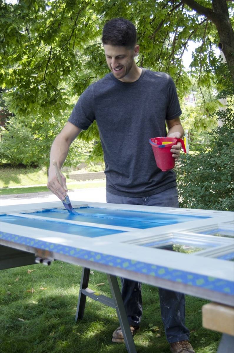 A man uses blue paint to fill in the interior panels of a door that's lying horizontally on a work table.