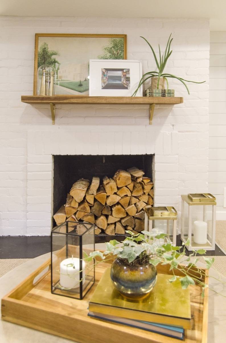 A fireplace stacked full of firewood on a white ship lapped wall, a wooden serving tray with a candle, potted plant and books, more candles by the fireplace.