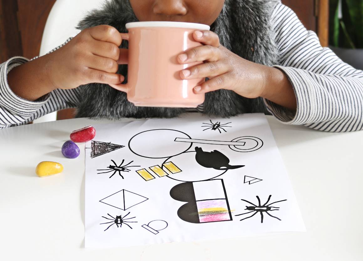 A girl drinks from a mug with a drawing in front of her on a white tabletop.