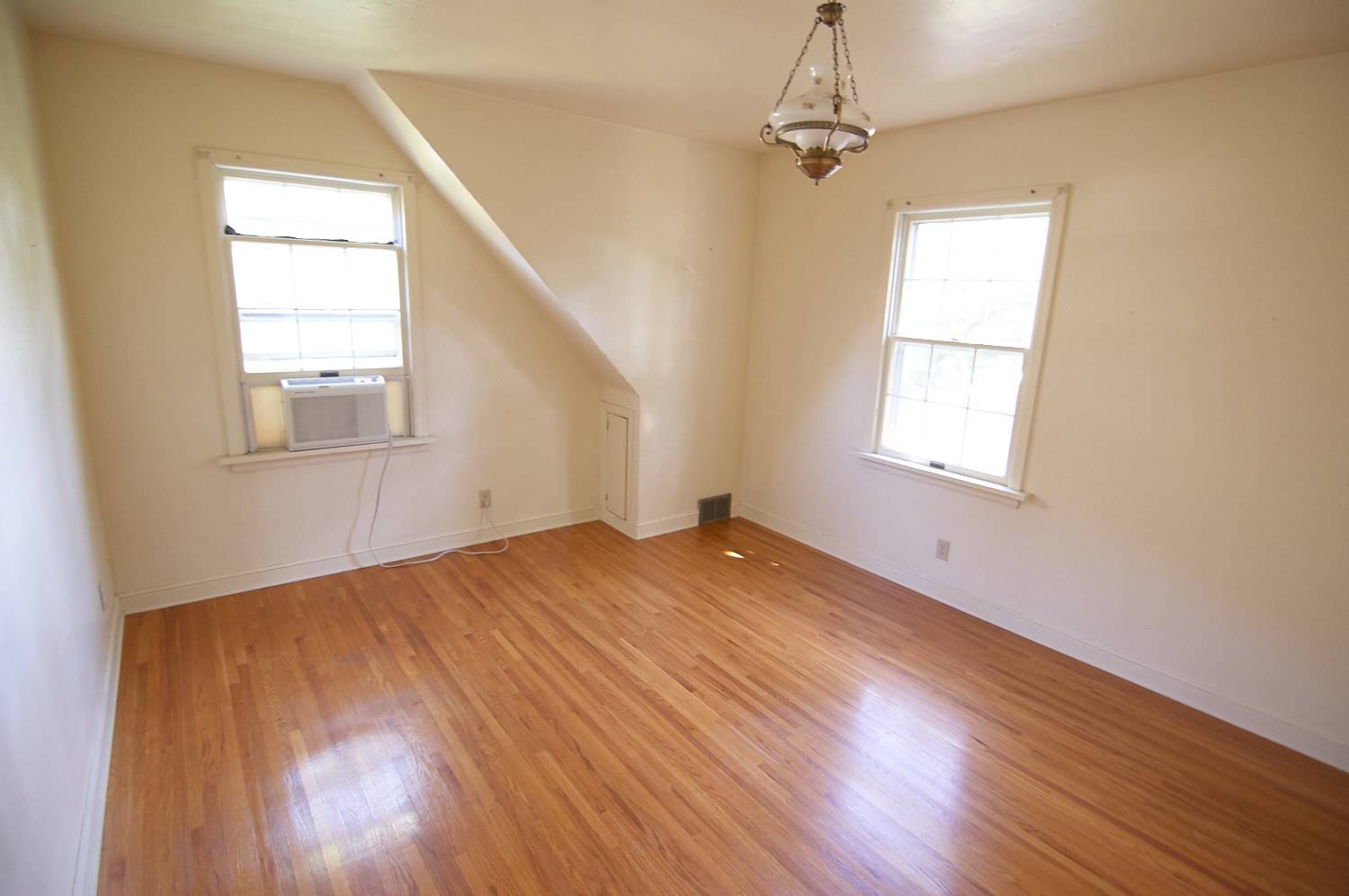 Interior of a room with two windows, white walls, hardwood floors, and a light fixture hanging from the ceiling.