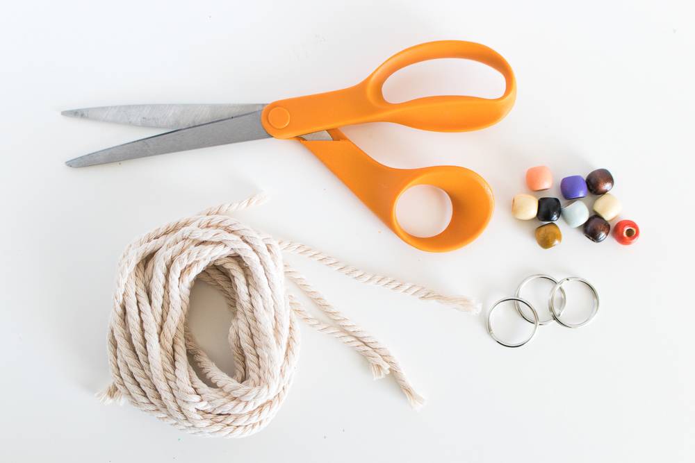 A pair of orange handles scissors, a roll of thin white rope, three metal rings and an assortment of colored beads.