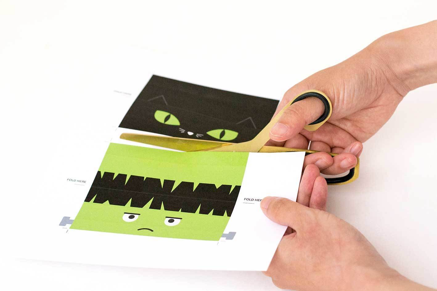 A woman uses scissors to cut two pieces of paper designed to look like Frankenstein and a black cat.