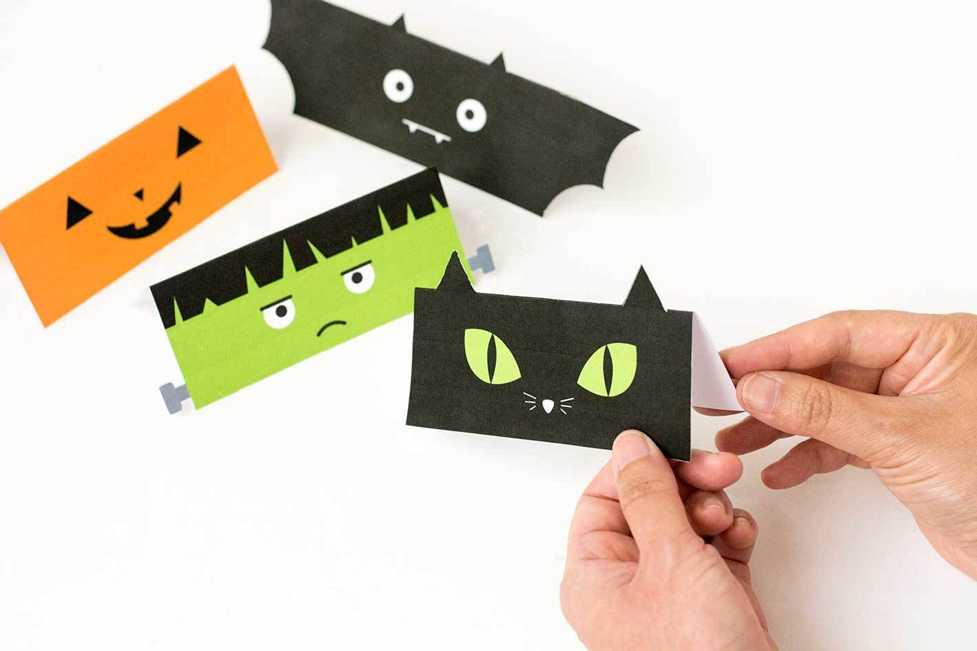 A lady is folding  paper, in half to look like a Jack-o-Lantern, black cats, Frankenstein and a bat to go on top of trick or treat bags.